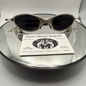 Peut inclure: Des lunettes de soleil argent&eacute;es avec des verres fonc&eacute;s reposent sur une surface r&eacute;fl&eacute;chissante. Elles sont pos&eacute;es au-dessus d'une carte de visite avec le texte "Paper Street Soap Co." et des coordonn&eacute;es, ainsi que "All Natural Handmade."