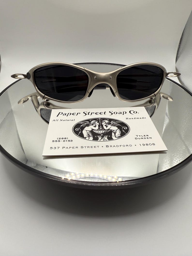 Peut inclure: Des lunettes de soleil argent&eacute;es avec des verres fonc&eacute;s reposent sur une surface r&eacute;fl&eacute;chissante. Elles sont pos&eacute;es au-dessus d'une carte de visite avec le texte "Paper Street Soap Co." et des coordonn&eacute;es, ainsi que "All Natural Handmade."
