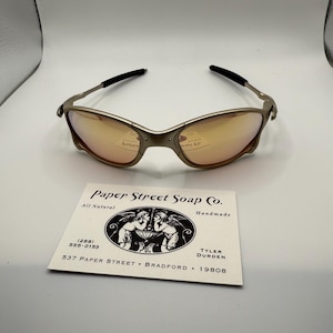 May include: A pair of gold-toned sunglasses with orange lenses and black arms. Below the sunglasses is a business card for Paper Street Soap Co., with the text "All Natural" and contact information.