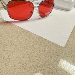 Puede incluir: Gafas de sol con montura plateada y lentes rojos. Las gafas est&aacute;n sobre una superficie blanca.