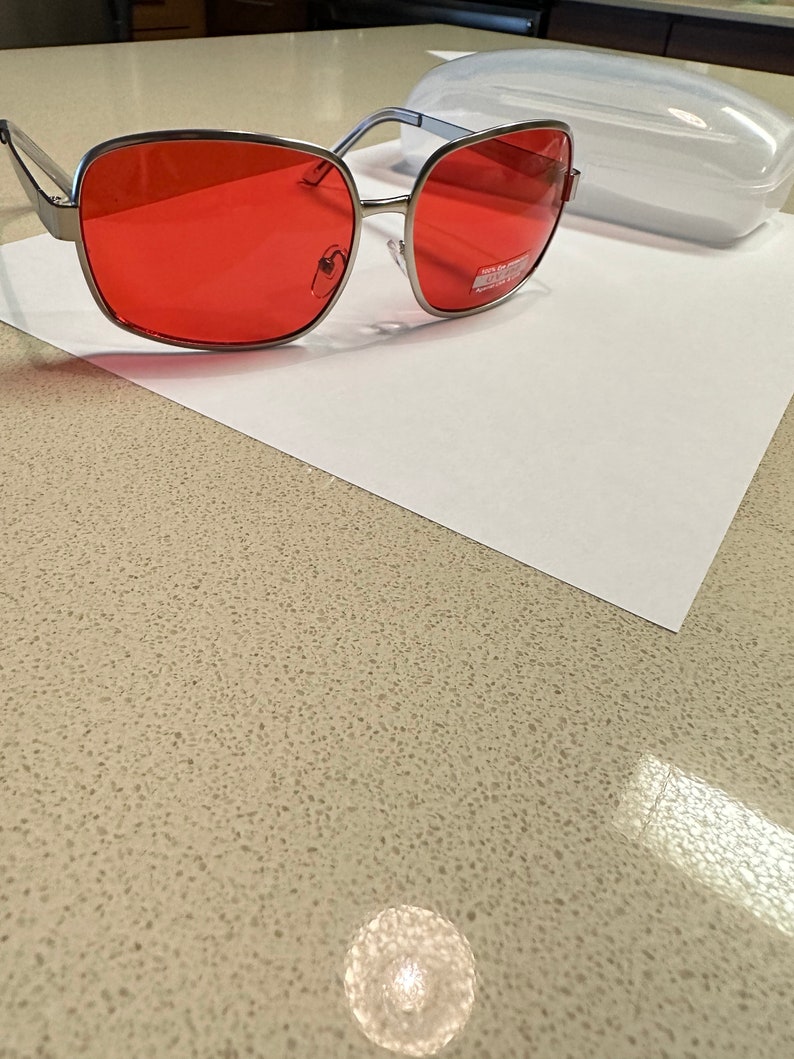 Puede incluir: Gafas de sol con montura plateada y lentes rojos. Las gafas est&aacute;n sobre una superficie blanca.