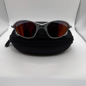 May include: Silver-framed sunglasses with orange-tinted lenses rest on a black zippered case. The sunglasses have a sleek, modern design with a metallic finish. The lenses reflect the surrounding environment.