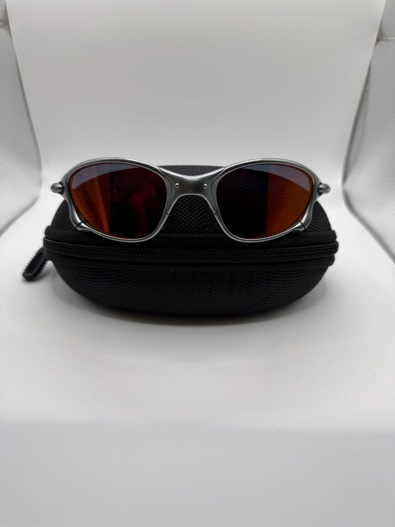 May include: Silver-framed sunglasses with orange-tinted lenses rest on a black zippered case. The sunglasses have a sleek, modern design with a metallic finish. The lenses reflect the surrounding environment.