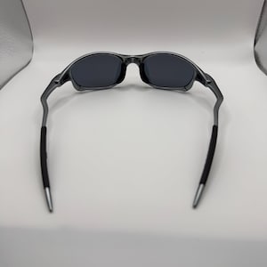 May include: Dark gray sunglasses with black lenses. The frames are a metallic gray, with black rubber grips on the temples. The tips of the temples are silver. The sunglasses are designed for a sporty look.