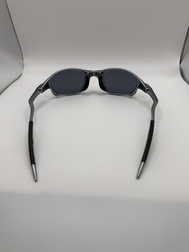 May include: Dark gray sunglasses with black lenses. The frames are a metallic gray, with black rubber grips on the temples. The tips of the temples are silver. The sunglasses are designed for a sporty look.