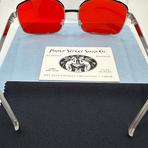 May include: Silver framed sunglasses with red lenses. The sunglasses are resting on a black fabric surface. The Paper Street Soap Co. business card is visible in the background.