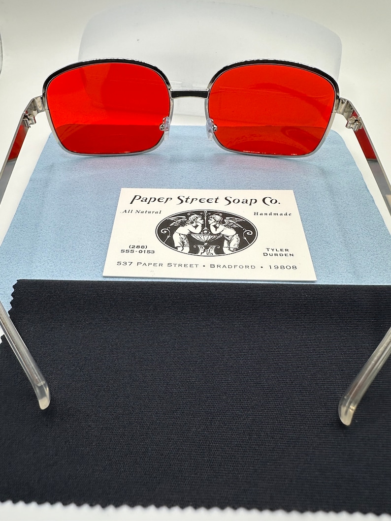 May include: Silver framed sunglasses with red lenses. The sunglasses are resting on a black fabric surface. The Paper Street Soap Co. business card is visible in the background.