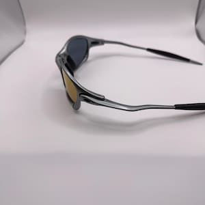 May include: A pair of silver-framed sunglasses with dark lenses and a gold-tinted lower lens. The temples are thin and extend to black tips. The sunglasses are set against a white background.
