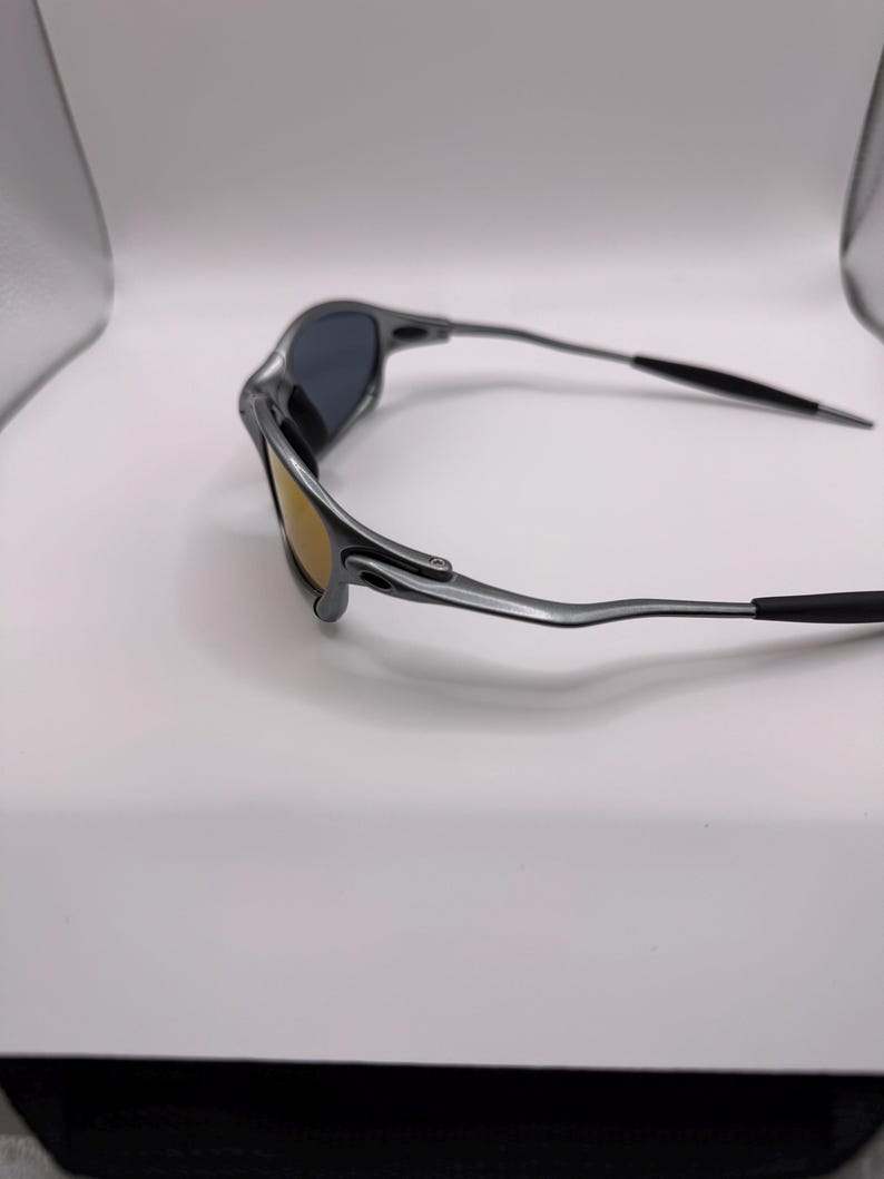 May include: A pair of silver-framed sunglasses with dark lenses and a gold-tinted lower lens. The temples are thin and extend to black tips. The sunglasses are set against a white background.