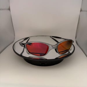 May include: Silver-framed sunglasses with red and orange lenses. The sunglasses are displayed on a reflective surface, showcasing their sleek design. The lenses have a gradient effect, transitioning from red to orange.