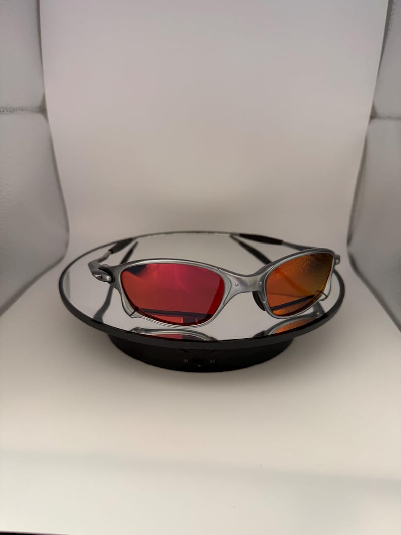 May include: Silver-framed sunglasses with red and orange lenses. The sunglasses are displayed on a reflective surface, showcasing their sleek design. The lenses have a gradient effect, transitioning from red to orange.
