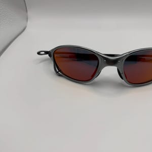 May include: Dark gray sunglasses with red-tinted lenses. The sunglasses have a sleek, modern design with a curved frame and a small, circular hinge detail. The lenses appear to be reflective, and the overall aesthetic is sporty.