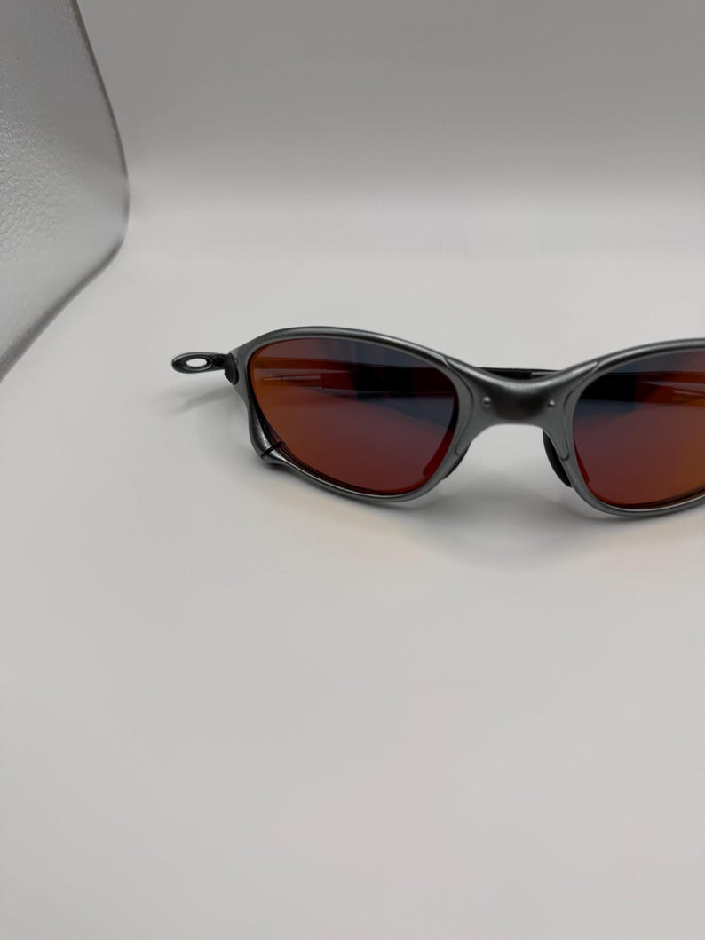 May include: Dark gray sunglasses with red-tinted lenses. The sunglasses have a sleek, modern design with a curved frame and a small, circular hinge detail. The lenses appear to be reflective, and the overall aesthetic is sporty.
