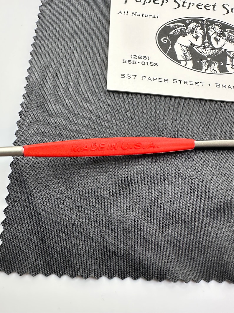 K&ouml;nnte beinhalten: Ein rotes Reinigungstuch mit einem Zickzackrand. Ein silbernes Reinigungswerkzeug mit einem roten "MADE IN USA." Etikett liegt auf dem Tuch. Das Tuch liegt auf einer wei&szlig;en Oberfl&auml;che. Eine wei&szlig;e Visitenkarte mit schwarzem Text ist im Hintergrund sichtbar. Der Text auf der Karte lautet "Paper Street So", "All Natural", "(288) 555-0153", und "537 Paper Street - Brat".