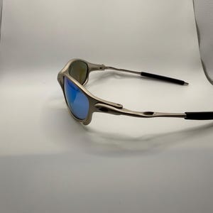 May include: A pair of silver-toned sunglasses with blue mirrored lenses. The sunglasses have a sleek, modern design with a thin frame and black earpieces. The lenses reflect a bright blue color.