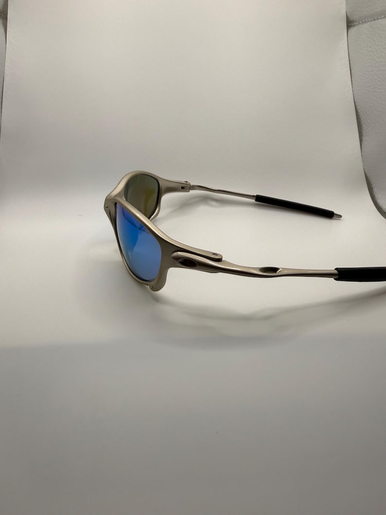 May include: A pair of silver-toned sunglasses with blue mirrored lenses. The sunglasses have a sleek, modern design with a thin frame and black earpieces. The lenses reflect a bright blue color.