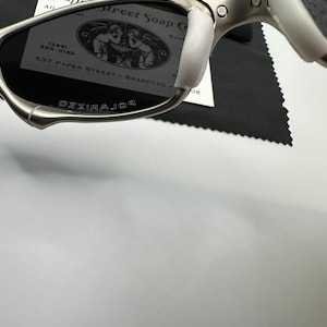 May include: Silver-framed sunglasses with dark lenses and white nose pads. The word "POLARIZED" is printed on the lens. A business card with the text "Paper Street Soap Co" is visible in the background.
