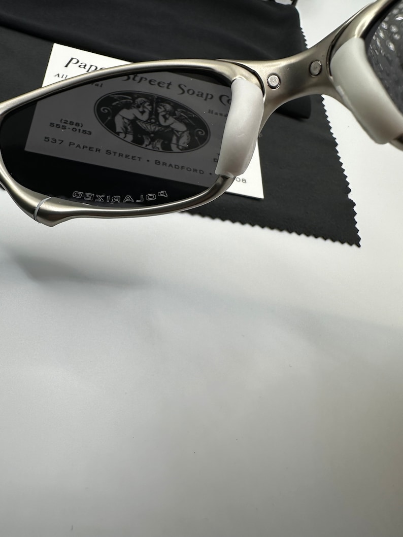 May include: Silver-framed sunglasses with dark lenses and white nose pads. The word "POLARIZED" is printed on the lens. A business card with the text "Paper Street Soap Co" is visible in the background.