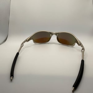 May include: Silver-framed sunglasses with orange-tinted lenses. The temples have black rubber sleeves for comfort. The sunglasses are designed for outdoor activities and offer protection from the sun.
