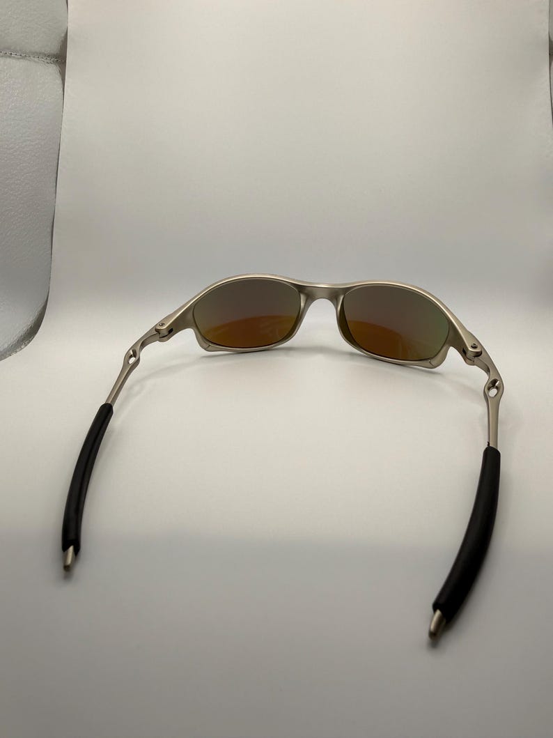 May include: Silver-framed sunglasses with orange-tinted lenses. The temples have black rubber sleeves for comfort. The sunglasses are designed for outdoor activities and offer protection from the sun.