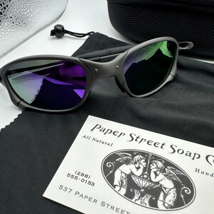 May include: Black sunglasses with a purple and green mirrored lens. The sunglasses are in a black case with a black microfiber cleaning cloth. The cleaning cloth has a business card on it that reads "Paper Street Soap Co. All Natural (288) 555-0153 537 Paper Street Bradford".
