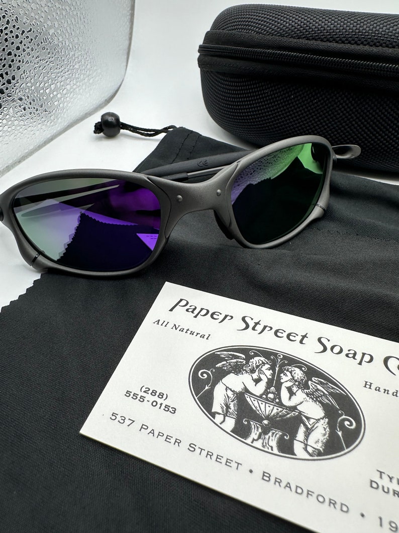 May include: Black sunglasses with a purple and green mirrored lens. The sunglasses are in a black case with a black microfiber cleaning cloth. The cleaning cloth has a business card on it that reads "Paper Street Soap Co. All Natural (288) 555-0153 537 Paper Street Bradford".