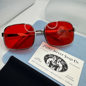 May include: Red square-shaped sunglasses with a silver frame. The sunglasses are sitting on a blue cleaning cloth and a business card for Paper Street Soap Co. The business card has a black and white illustration of two angels holding a bowl. The text on the business card reads "Paper Street Soap Co. All Natural Handmade (288) 555-0153 537 Paper Street, Bradford, Tyler Durden 19808."