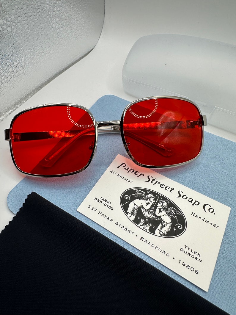 May include: Red square-shaped sunglasses with a silver frame. The sunglasses are sitting on a blue cleaning cloth and a business card for Paper Street Soap Co. The business card has a black and white illustration of two angels holding a bowl. The text on the business card reads "Paper Street Soap Co. All Natural Handmade (288) 555-0153 537 Paper Street, Bradford, Tyler Durden 19808."