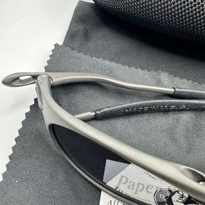 May include: A pair of silver-toned sunglasses with dark lenses. The sunglasses are resting on a gray fabric surface. The sunglasses have a metal frame and a black leather strap. The text "MADE IN U.S.A." is visible on the strap. The sunglasses are resting on a white card with the text "Pap&eacute;" and a logo.