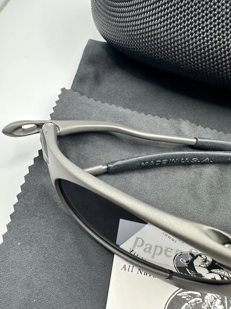 May include: A pair of silver-toned sunglasses with dark lenses. The sunglasses are resting on a gray fabric surface. The sunglasses have a metal frame and a black leather strap. The text "MADE IN U.S.A." is visible on the strap. The sunglasses are resting on a white card with the text "Pap&eacute;" and a logo.