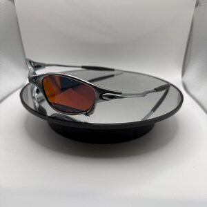 May include: Silver-framed sunglasses with red-tinted lenses. The sunglasses are displayed on a reflective surface, showcasing their sleek design. The temples are thin and curve around the ears. The overall aesthetic is modern and sporty.