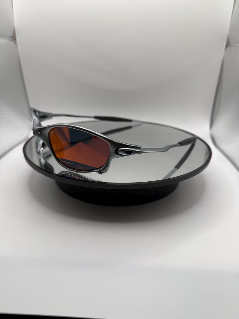 May include: Silver-framed sunglasses with red-tinted lenses. The sunglasses are displayed on a reflective surface, showcasing their sleek design. The temples are thin and curve around the ears. The overall aesthetic is modern and sporty.