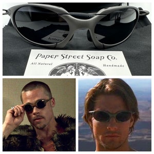 May include: A close-up of a pair of black sunglasses with a wraparound design. The sunglasses are sitting on a black fabric surface. The text "Paper Street Soap Co. All Natural Handmade" is visible in the image.