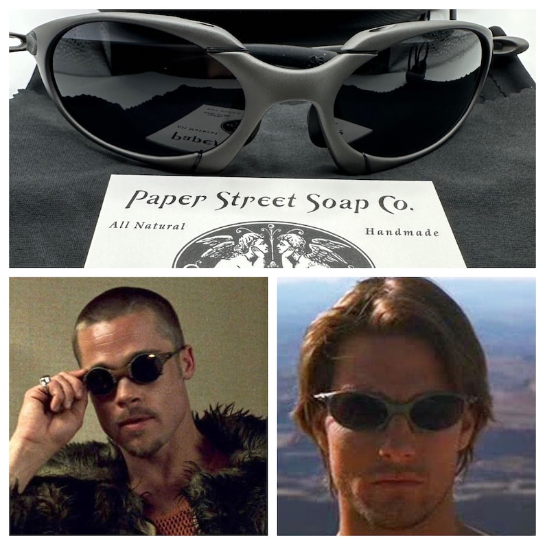 May include: A close-up of a pair of black sunglasses with a wraparound design. The sunglasses are sitting on a black fabric surface. The text "Paper Street Soap Co. All Natural Handmade" is visible in the image.