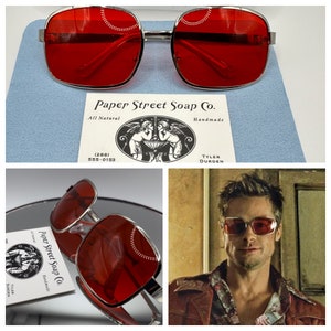 May include: Red square-shaped sunglasses with a silver frame. The sunglasses are on a blue surface with a business card that reads "Paper Street Soap Co. All Natural Handmade (288) 555-0153 TYLER DURDEN".