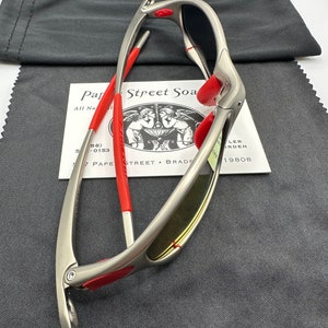 K&ouml;nnte beinhalten: Eine silberfarbene Sonnenbrille mit roten Akzenten und gelbt&ouml;nenden Gl&auml;sern. Die Sonnenbrille ist zusammengeklappt und liegt auf einem grauen Mikrofaser-Reinigungstuch. Das Tuch tr&auml;gt den Text "Par Street Soa".