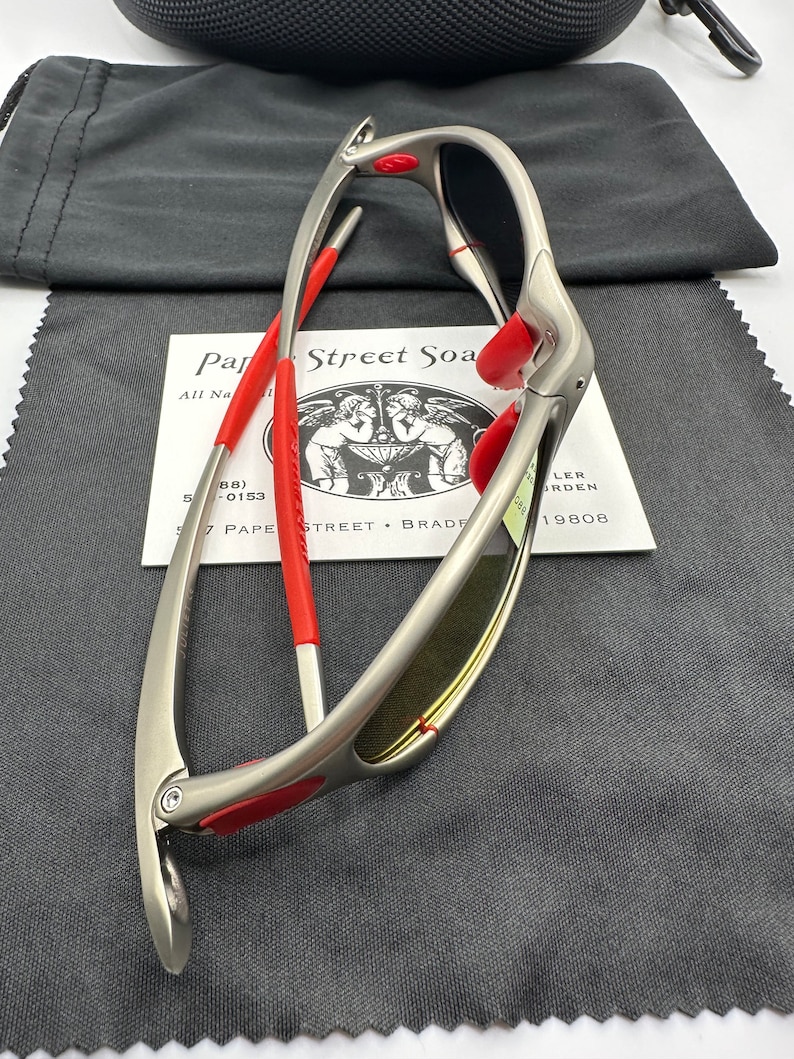 K&ouml;nnte beinhalten: Eine silberfarbene Sonnenbrille mit roten Akzenten und gelbt&ouml;nenden Gl&auml;sern. Die Sonnenbrille ist zusammengeklappt und liegt auf einem grauen Mikrofaser-Reinigungstuch. Das Tuch tr&auml;gt den Text "Par Street Soa".