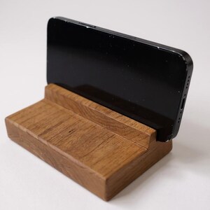 May include: A wooden phone stand with a slot to hold a smartphone. The stand is made of light brown wood and has a rectangular base. A black smartphone is placed in the stand.