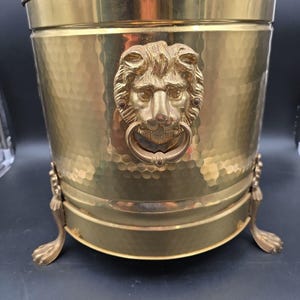 Puede incluir: Un cubo cilíndrico de latón con textura martillada y acabado pulido. El exterior está adornado con un relieve prominente de cabeza de león que sostiene un anillo en la boca. El recipiente se apoya sobre tres patas ornamentadas con forma de garra.