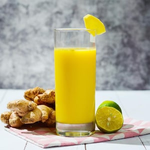 May include: A tall glass filled with a vibrant yellow beverage, garnished with a lemon wedge. Fresh ginger roots and a halved lime rest on a pink and white checkered napkin, suggesting a refreshing drink.