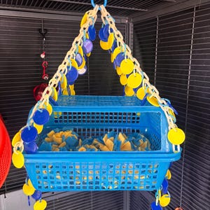 May include: A blue plastic pet cage hanging basket filled with blue and yellow fabric pieces. The basket is suspended by a chain of yellow and white plastic links with blue and yellow circular accents.