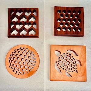 May include: Four wooden coasters with different designs. The first coaster features a pattern of hearts, the second has a honeycomb pattern, the third has a circle pattern, and the fourth has a turtle design.