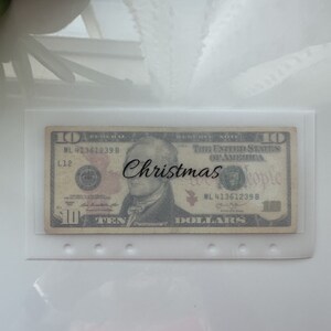 May include: A ten dollar bill with the text "Christmas" written on it. The bill is in a clear plastic sleeve with holes punched in the top.