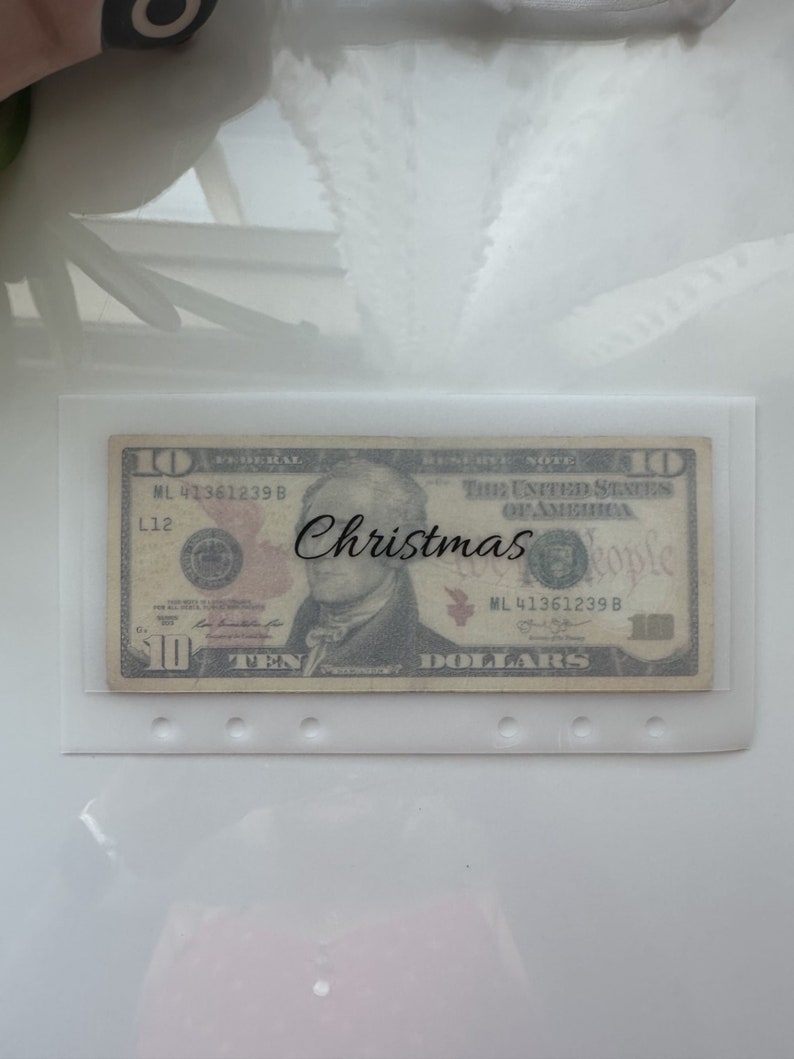 May include: A ten dollar bill with the text "Christmas" written on it. The bill is in a clear plastic sleeve with holes punched in the top.