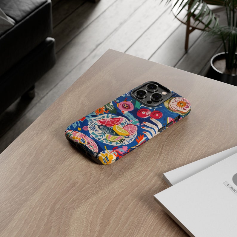 May include: A colorful phone case with a vibrant floral and fruit design. The case features a dark blue background with illustrations of lemons, cherries, and flowers. The case is on a light wood surface.