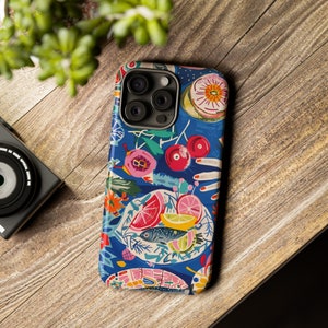 May include: A blue iPhone case with a colorful, artistic design featuring food illustrations. The design includes images of cherries, fish, citrus fruits, and flowers. The case is on a wooden surface.
