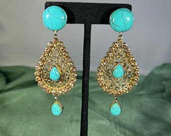 May include: A pair of gold dangle earrings with turquoise stones. The earrings feature a circular top with a smaller turquoise stone hanging below. The earrings are on a black stand.