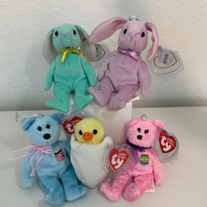 May include: A collection of five plush Easter-themed beanie babies. The beanie babies include a light blue bear, a pink bear, a light green bunny, a purple bunny, and a yellow chick in a white egg.