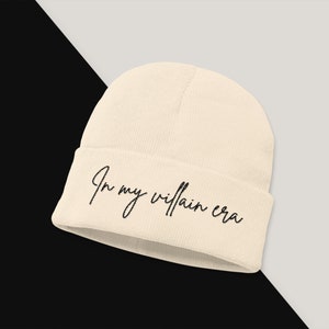 May include: A beige beanie with the text "In my villain era" embroidered in black.