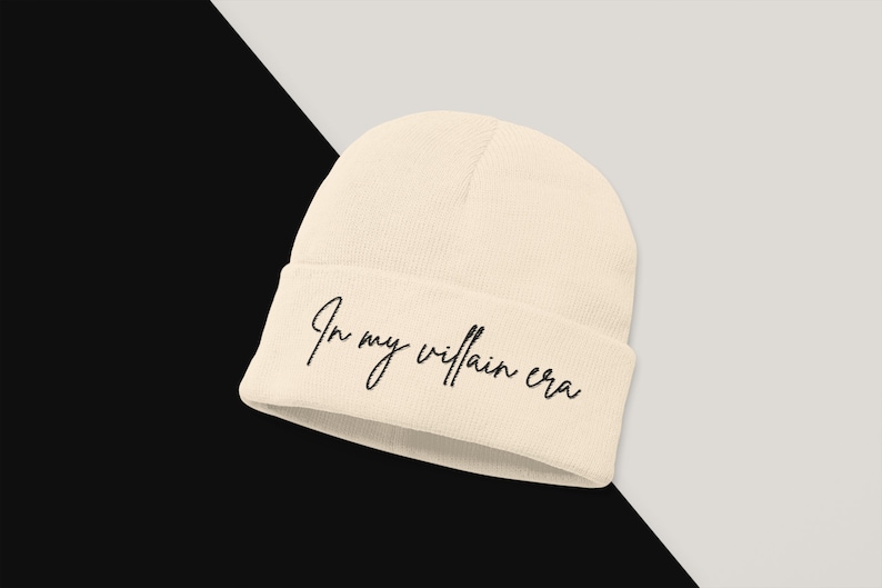 May include: A beige beanie with the text "In my villain era" embroidered in black.